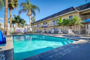 A swimming pool with lounge chairs and umbrellas, surrounded by palm trees and a building with balconies.