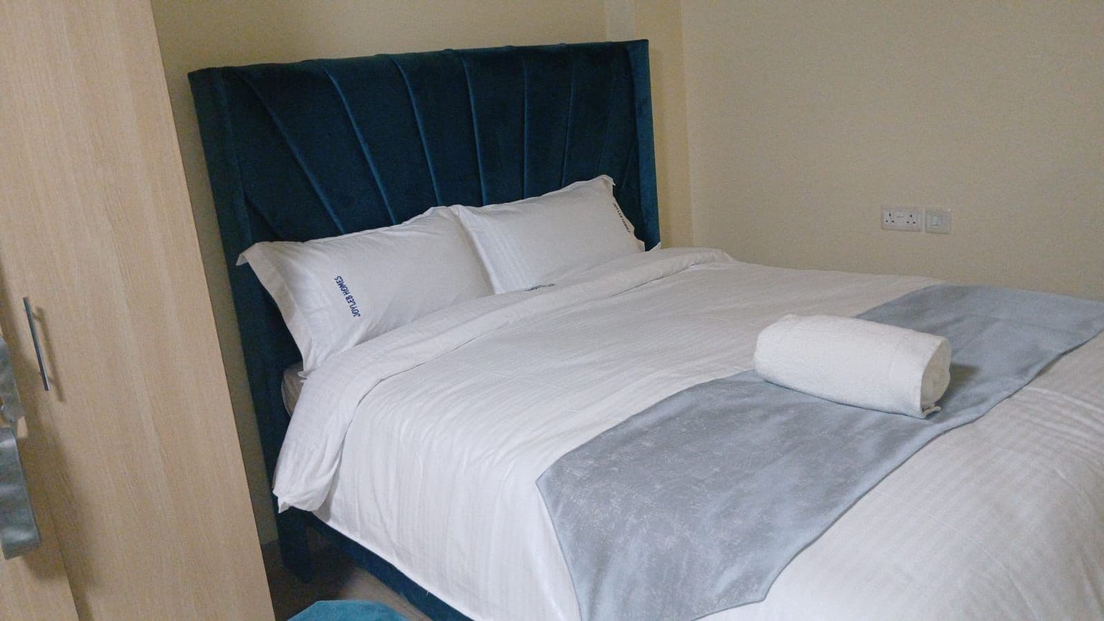 Una cama con cabecera oscura, ropa de cama blanca y una toalla enrollada.