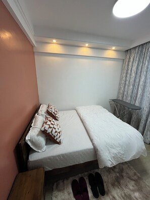 A bedroom with a bed, pillows, a small table, and a chair. - JKIA Terminal Luxore  (Syokimau)