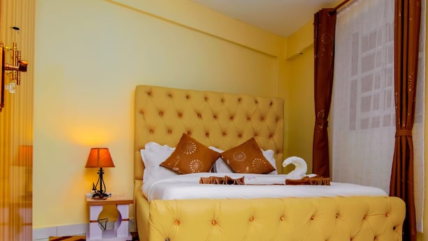 A hotel room with a large bed, a nightstand with a lamp, a patterned rug, and a window with curtains. - JKIA Terminal Luxore  (Syokimau)