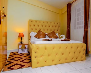 A hotel room with a large bed, a nightstand with a lamp, a patterned rug, and a window with curtains. - JKIA Terminal Luxore  (Syokimau)