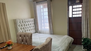 A bedroom with a bed, a wooden table, two chairs, a potted plant, and a door with windows. - JKIA Terminal Luxore  (Syokimau)