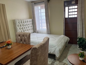A bedroom with a bed, a wooden table, two chairs, a potted plant, and a door with windows. - JKIA Terminal Luxore  (Syokimau)