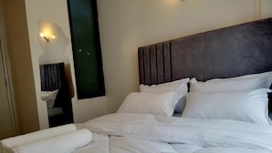 A neatly made bed with white linens and pillows, a dark headboard, and a wall-mounted light fixture. - JKIA Terminal Luxore  (Syokimau)