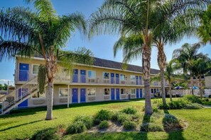 Un hotel con una estructura de dos pisos, varias habitaciones con puertas azules, una escalera central y un césped bien cuidado con palmeras.