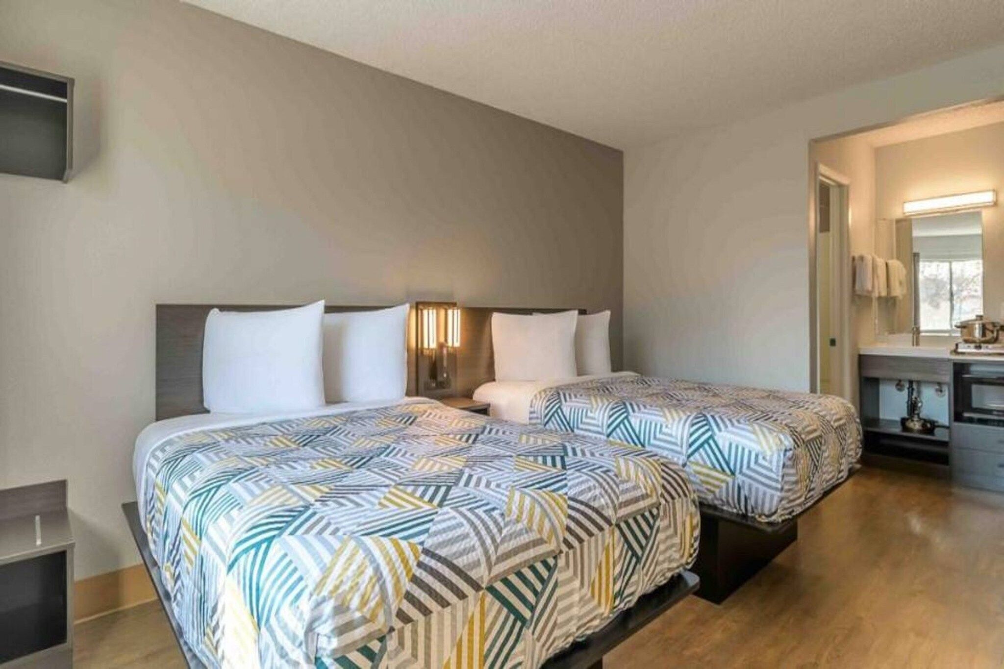 A hotel room with two beds, a bedside table, a mirror, and a kitchenette.