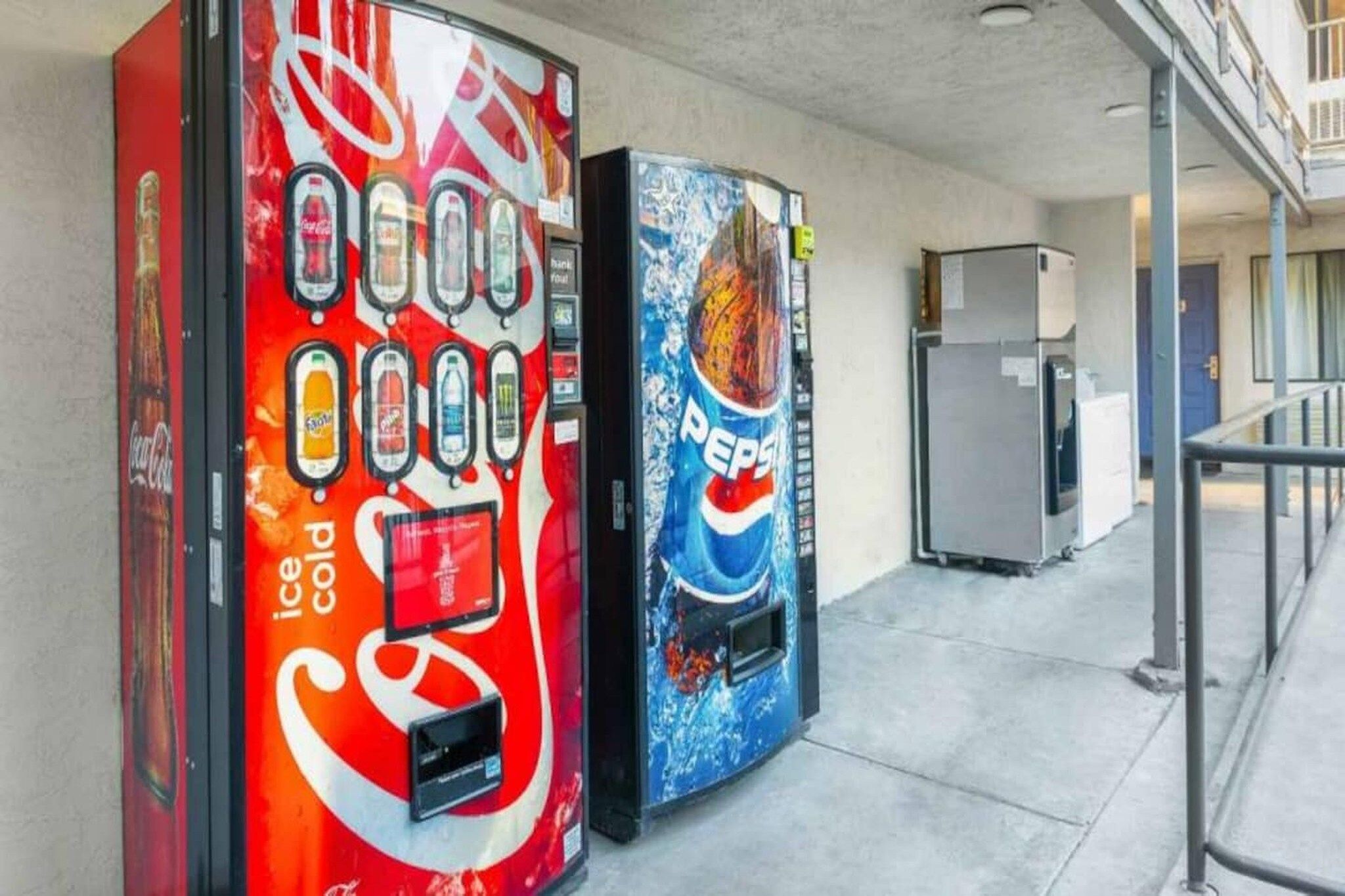 Two vending machines, one with a Coca-Cola logo and the other with a Pepsi logo, in a corridor with a refrigerator in the background.