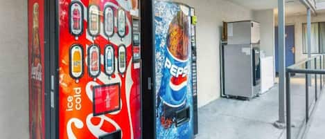 Two vending machines, one with a Coca-Cola logo and the other with a Pepsi logo, in a corridor with a refrigerator in the background.