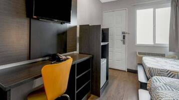 Habitación de hotel con escritorio, silla, televisor, armario y cama con ropa de cama estampada.
