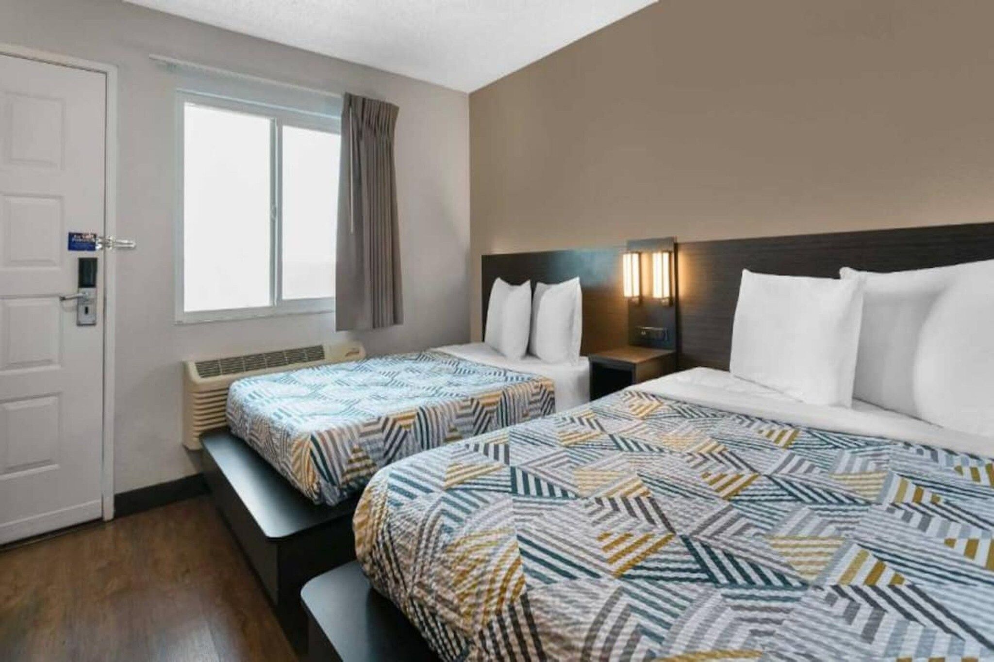 A hotel room with two beds, a window with curtains, a bedside table with a lamp, and a door with a lock.