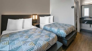 A hotel room with two beds, a bedside table with a lamp, a mirror, and a bathroom with a sink and mirror. - Sunny 2 Full Beds | Smoking, Micfridge (Merced)