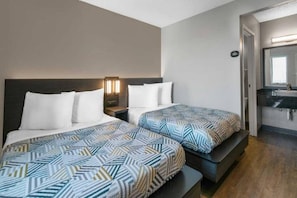 Quarto de hotel com duas camas, mesa de cabeceira com luminária, espelho e banheiro com pia e espelho.