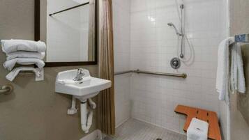 Een standaard hotelbadkamer met een wastafel, spiegel, douche en een klein houten bankje.
