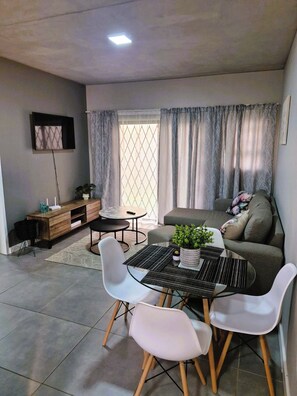 Ein modernes Wohnzimmer mit einer Couch, einem Esstisch mit Stühlen, einem an der Wand befestigten Fernseher und einem Fenster mit Vorhängen.