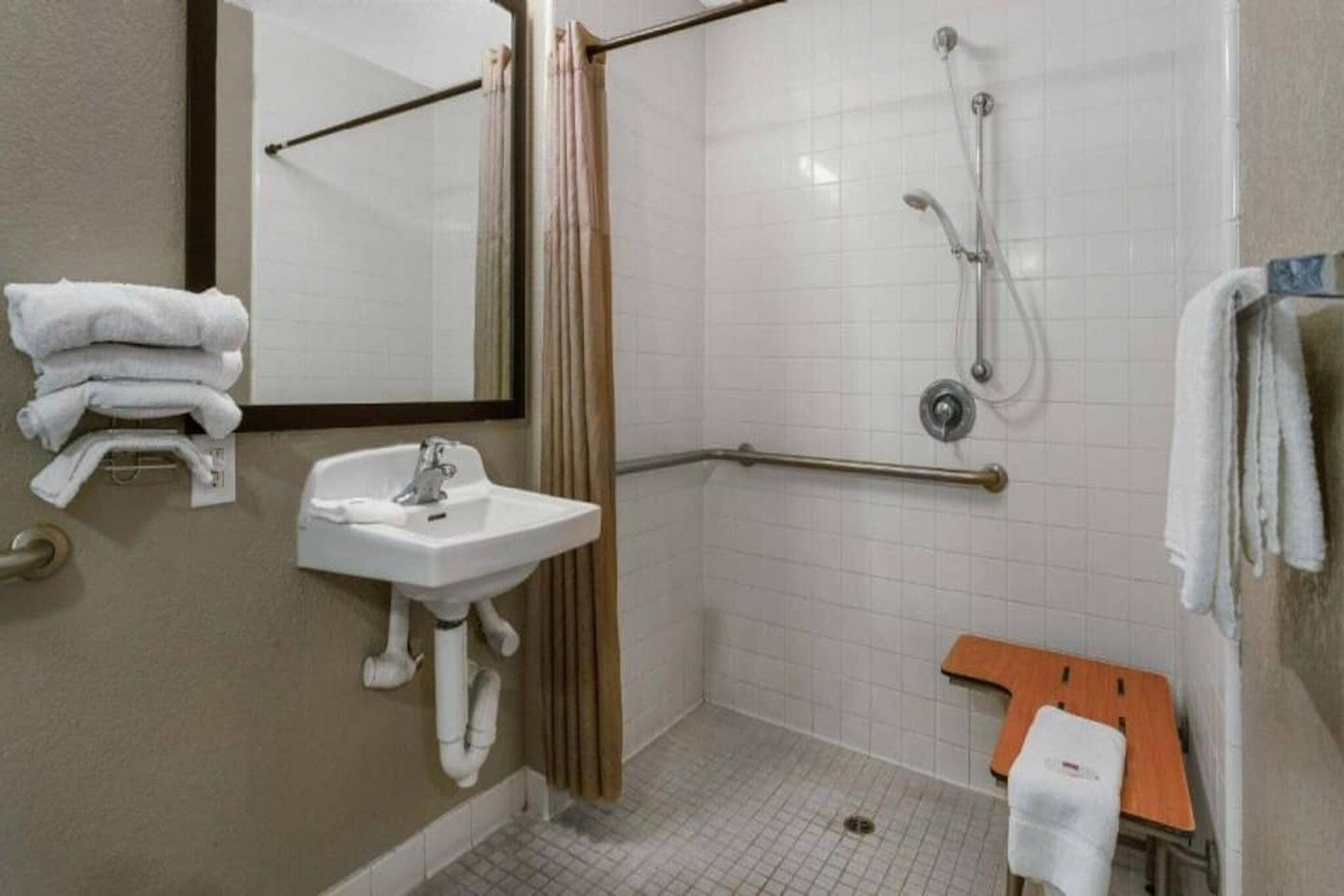 A standard hotel bathroom with a sink, mirror, shower, and a small wooden bench.