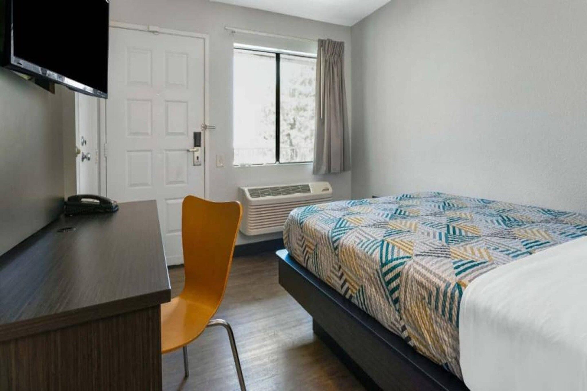 A bedroom with a bed, a desk, a chair, a TV, and a window.