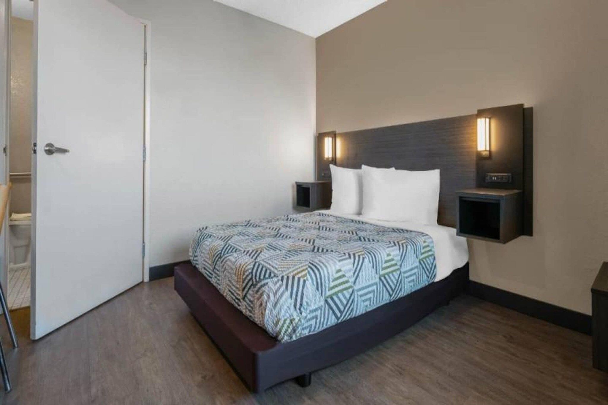 A modern hotel room with a large bed, a bedside table, a nightstand, and a wall-mounted light fixture.