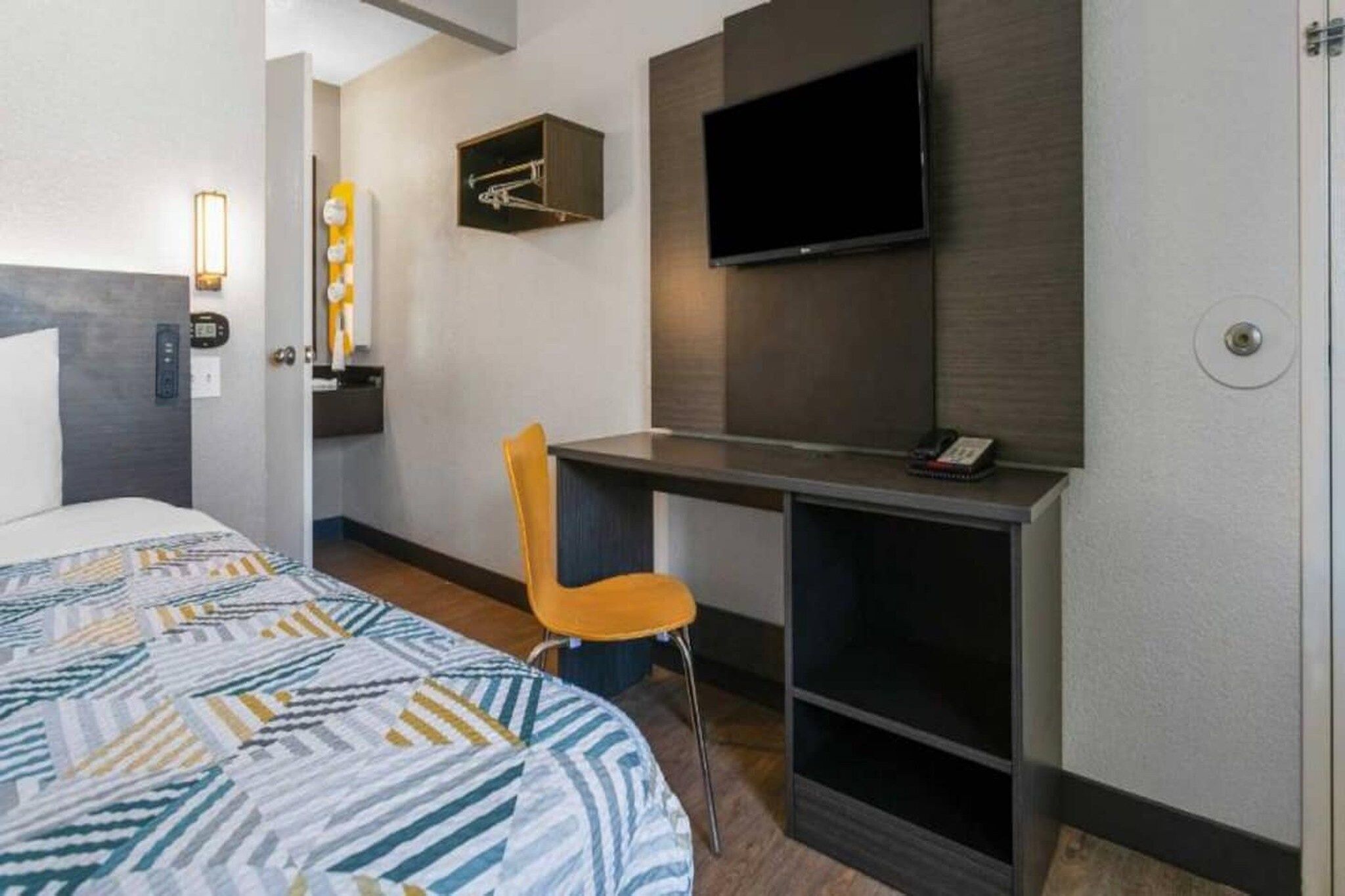 A hotel room with a bed, a desk, a chair, a television, and a wall-mounted light fixture.