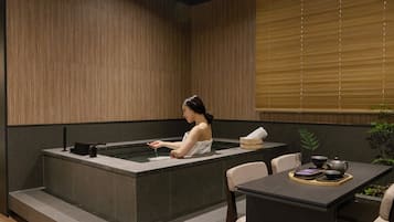 A spa room with a hot tub, a table with tea set, and a window with blinds.