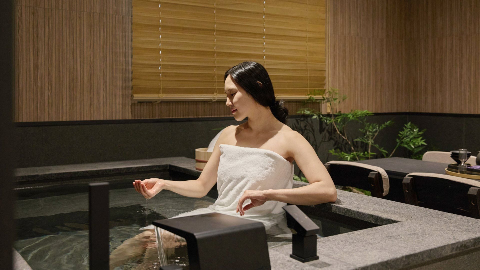 A person sitting in a hot spring with a towel wrapped around their body, a stone bench, and a table with a glass and a book.