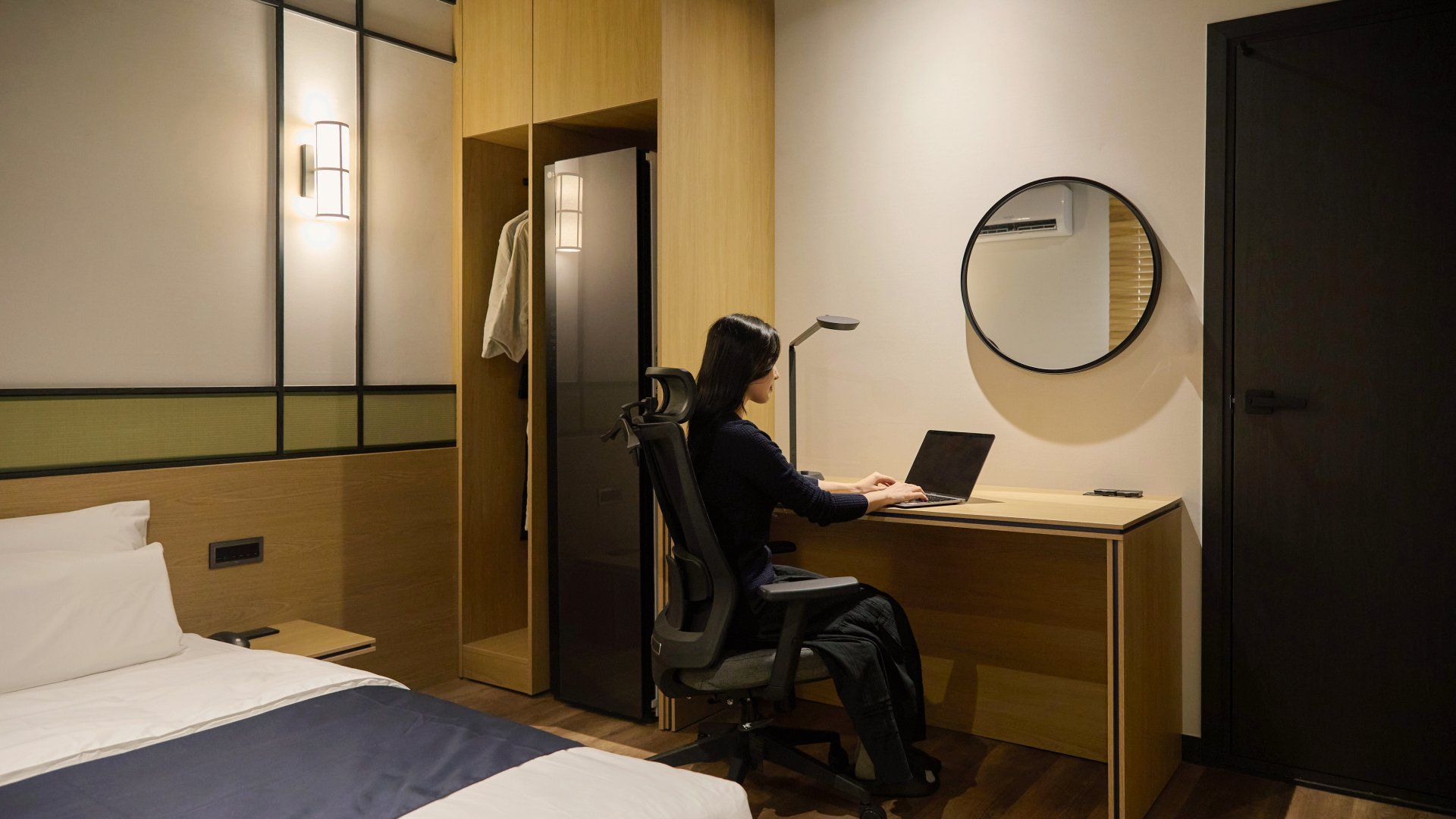 A hotel room with a bed, a desk with a laptop, a chair, a wardrobe, and a mirror.