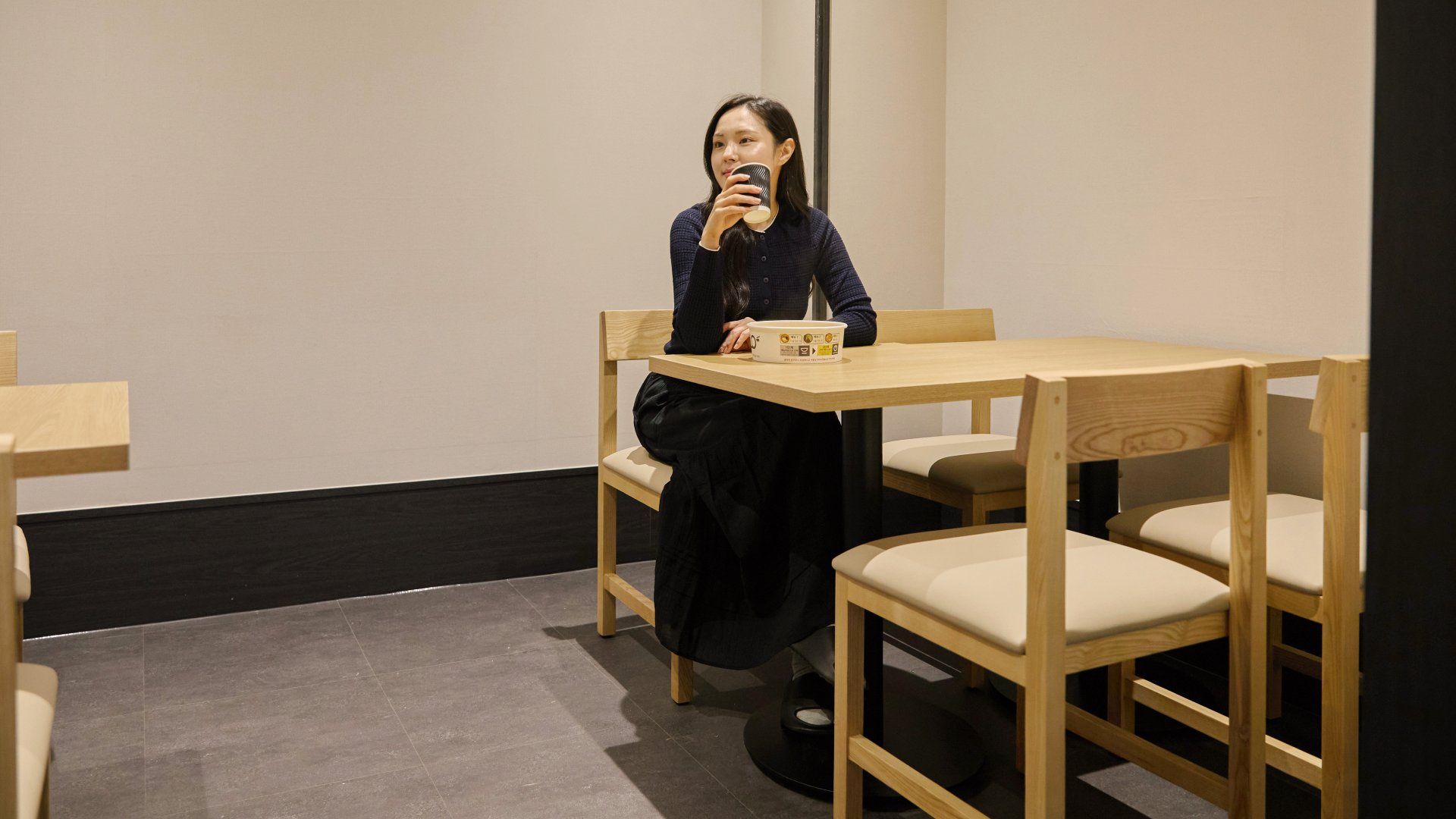Eine Person sitzt an einem Holztisch mit einer Tasse in einem Raum mit schlichten Wänden und Fliesenboden.