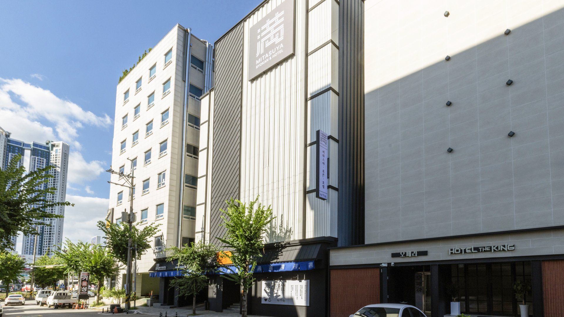 A modern hotel building with a prominent sign for 'Mita Suya Ryokan & Hotel' and a smaller sign for 'Hotel The King' across the street.