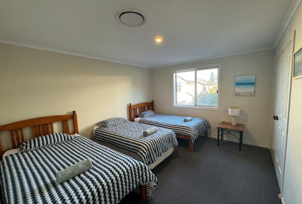 A room with three single beds, a small table with a lamp, and a window with a view of buildings.