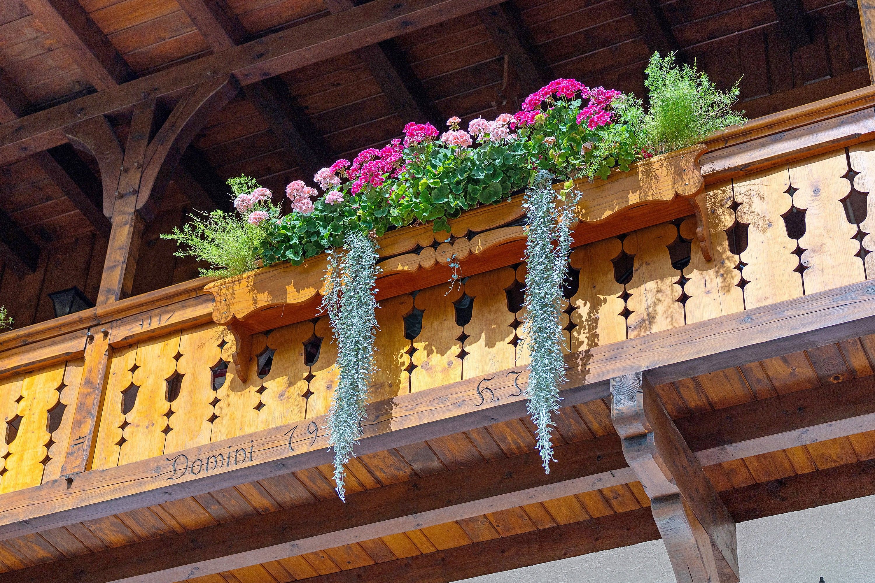 A wooden balcony with a wrought iron railing, a wooden sign with the name 'Domini', and hanging flower baskets with pink flowers and green foliage.