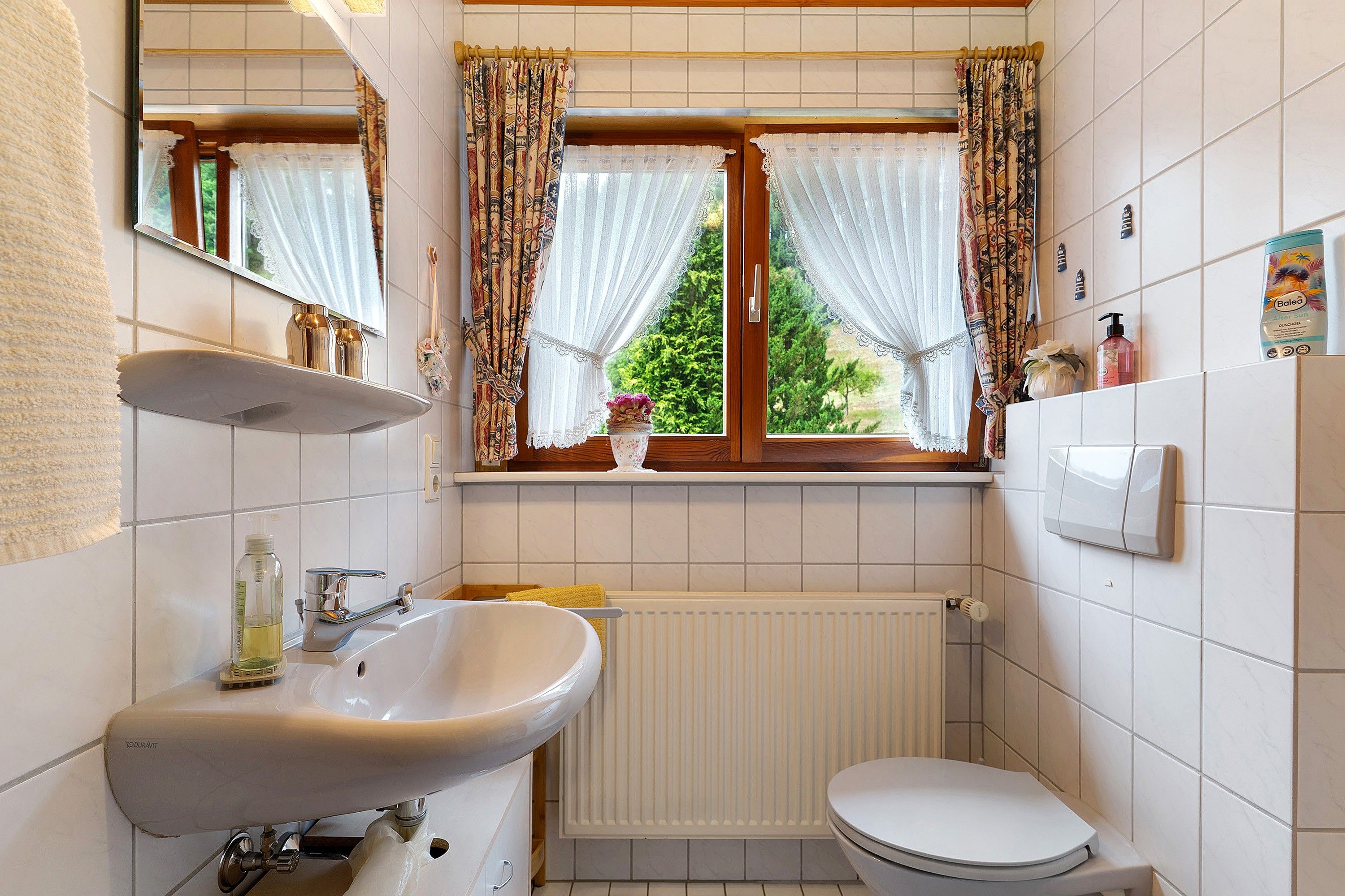 Ein Badezimmer mit einem weißen Waschbecken, einer Toilette, einem Fenster mit gemusterten Vorhängen und einem Heizkörper.