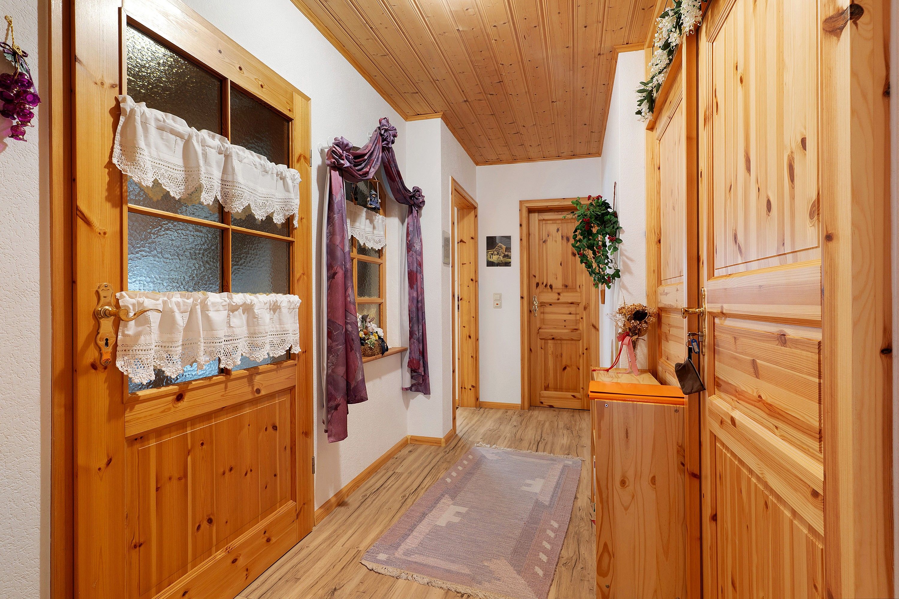 A hallway with wooden doors, a window with lace curtains, and a small shelf with a plant.