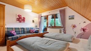 Ein gemütliches Zimmer mit Holzbalkendecke, einer Couch mit Karokissen, einem Bett mit weißer Tagesdecke und einem Fenster mit Vorhängen.