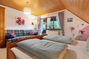 Una habitación acogedora con techo de madera, un sofá con cojines escoceses, una cama con colcha blanca y una ventana con cortinas.