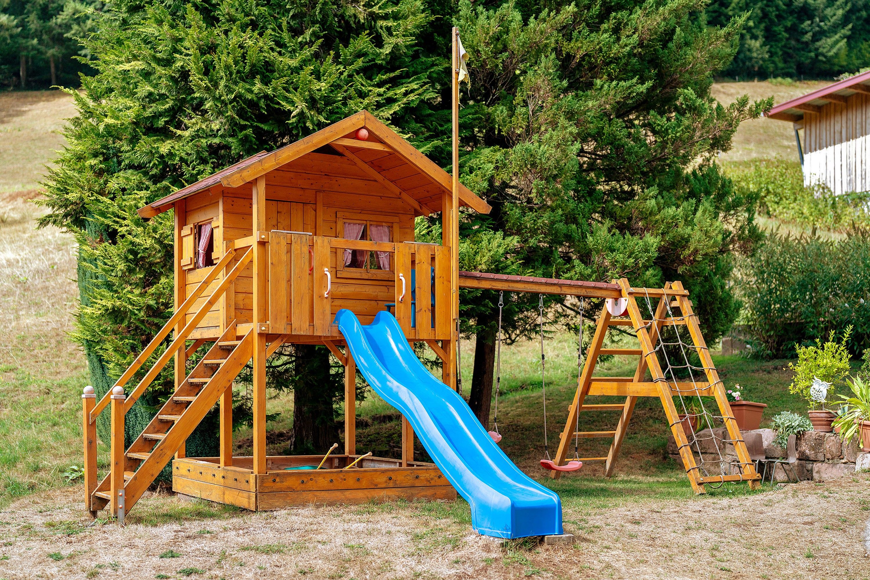 A wooden playhouse with a slide and climbing area, surrounded by trees and grass.