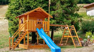 A wooden playhouse with a slide and climbing area, surrounded by trees and grass.