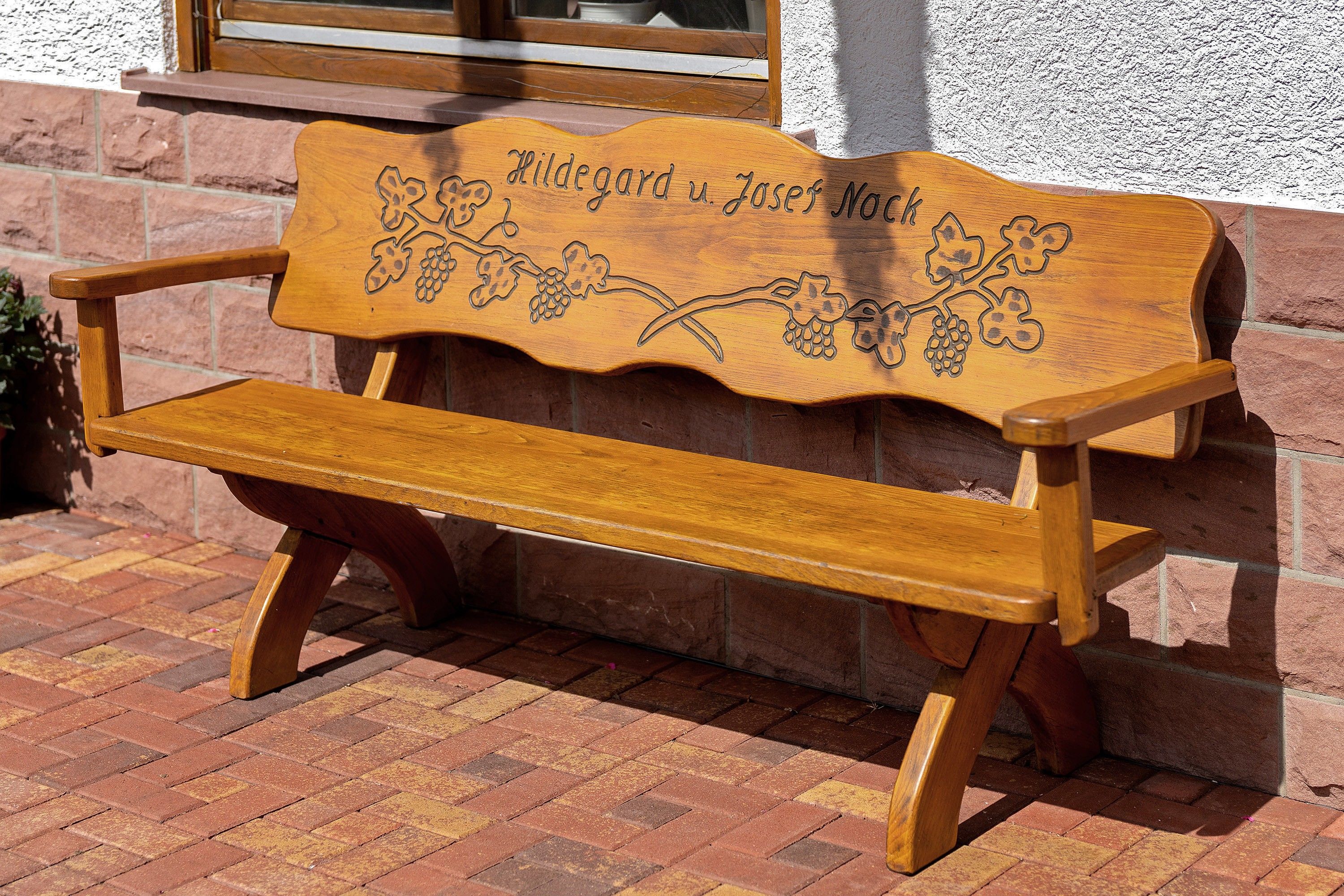 A wooden bench with a carved design and text, placed against a brick wall.