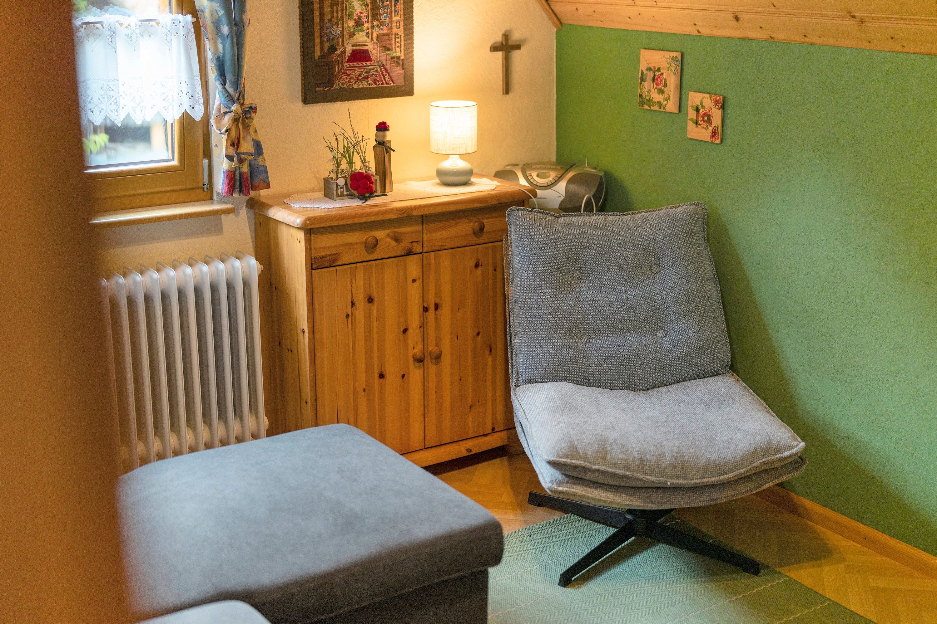 A cozy room with a wooden cabinet, a cushioned bench, a chair with a patterned cushion, a radiator, and a wall with a cross and two framed pictures.