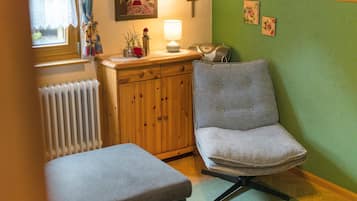 A cozy room with a wooden cabinet, a cushioned bench, a chair with a patterned cushion, a radiator, and a wall with a cross and two framed pictures.