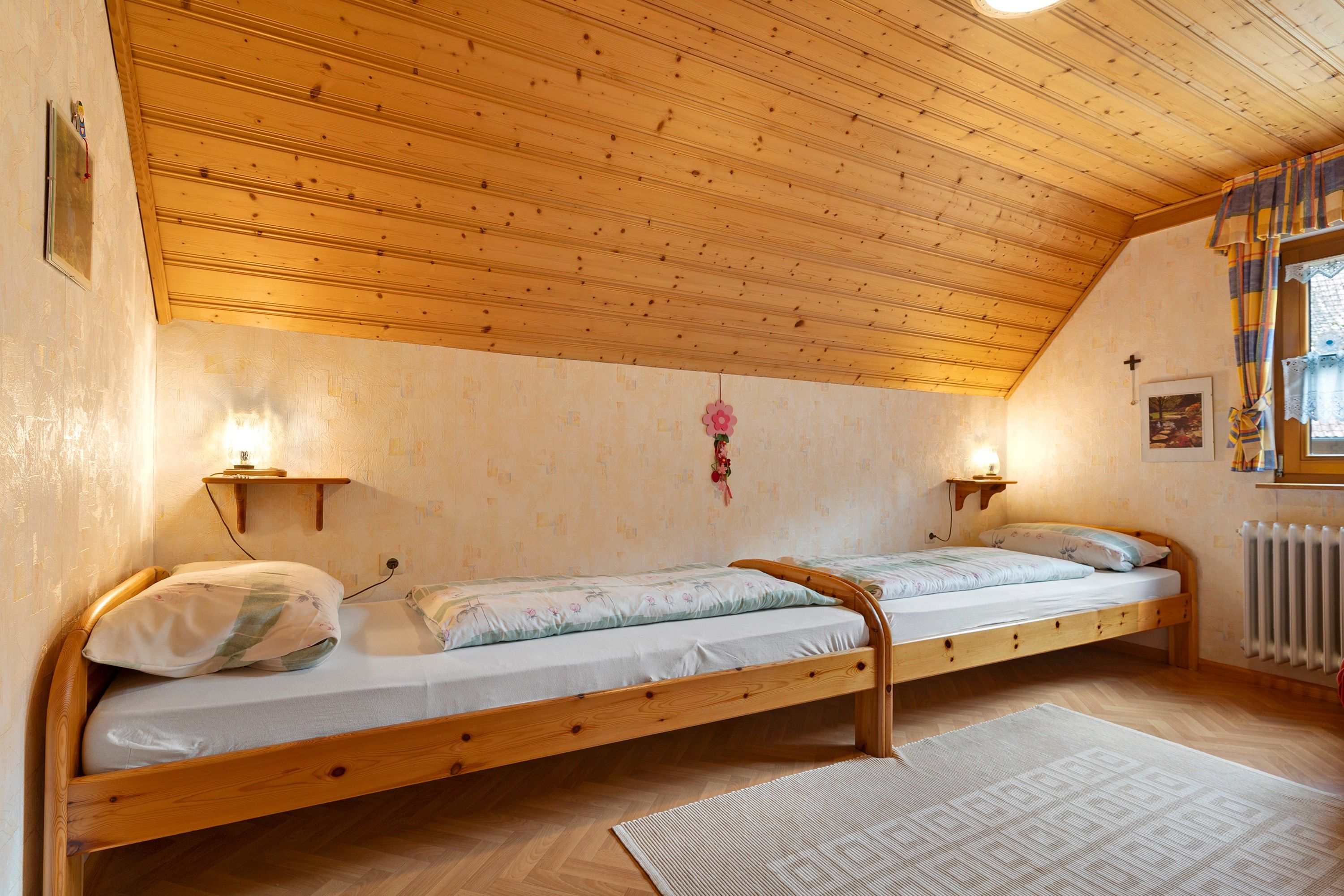 A room with two wooden bunk beds, a wooden ceiling, a radiator, and a window with curtains.