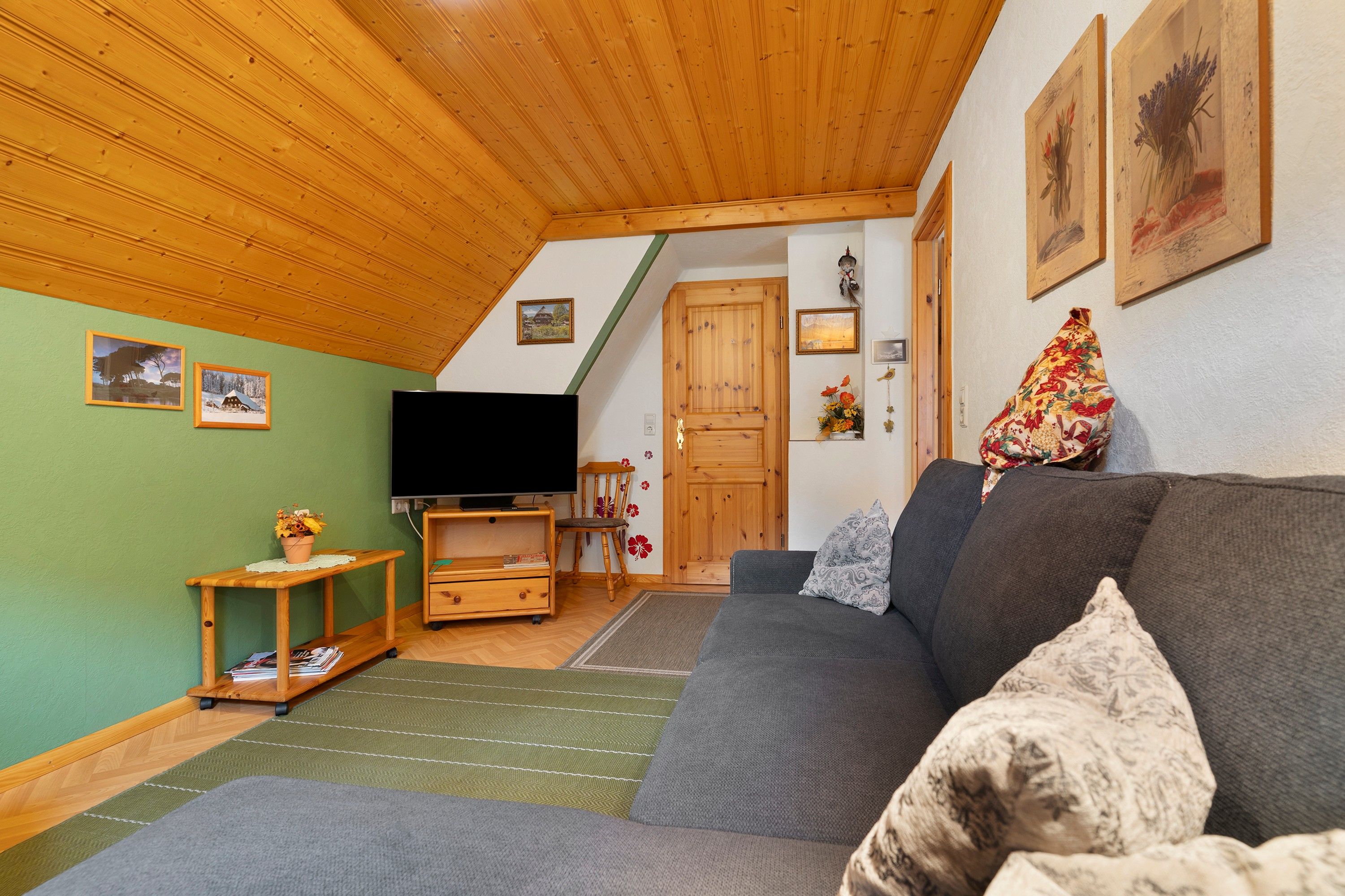 Ein gemütliches Wohnzimmer mit Holzbalkendecke, einem grauen Sofa, einem Fernseher und einem kleinen Holztisch mit einer Pflanze.