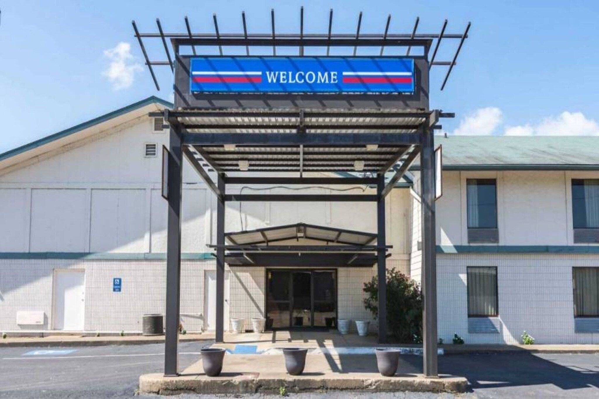 Entrada de um edifício com uma placa de 'WELCOME', área de espera coberta e estacionamento.