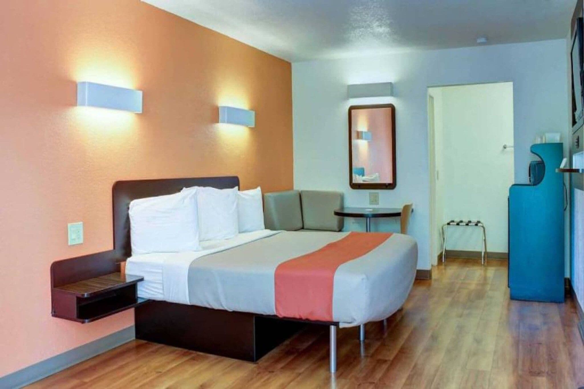 A hotel room with a bed, bedside table, a small table with a chair, a mirror, and a kitchenette area.
