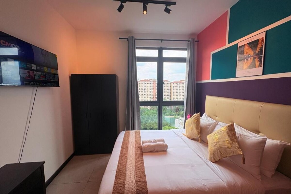 A hotel room with a bed, a TV on the wall, a window with curtains, and a view of buildings outside.