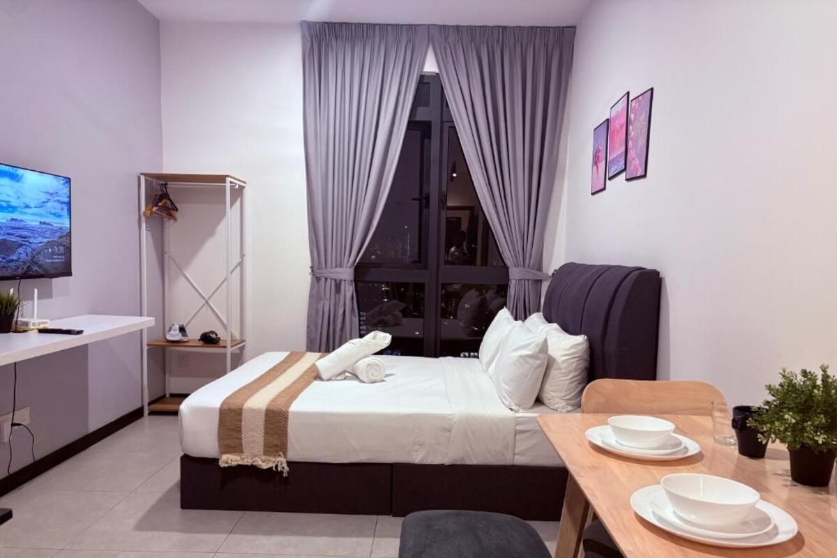 A modern hotel room with a bed, bedside table, TV, and a dining area with a table and chairs.