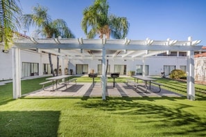 Un espace extérieur abrité, doté de tables et de chaises, entouré d’une pergola blanche et agrémenté de palmiers en arrière-plan.