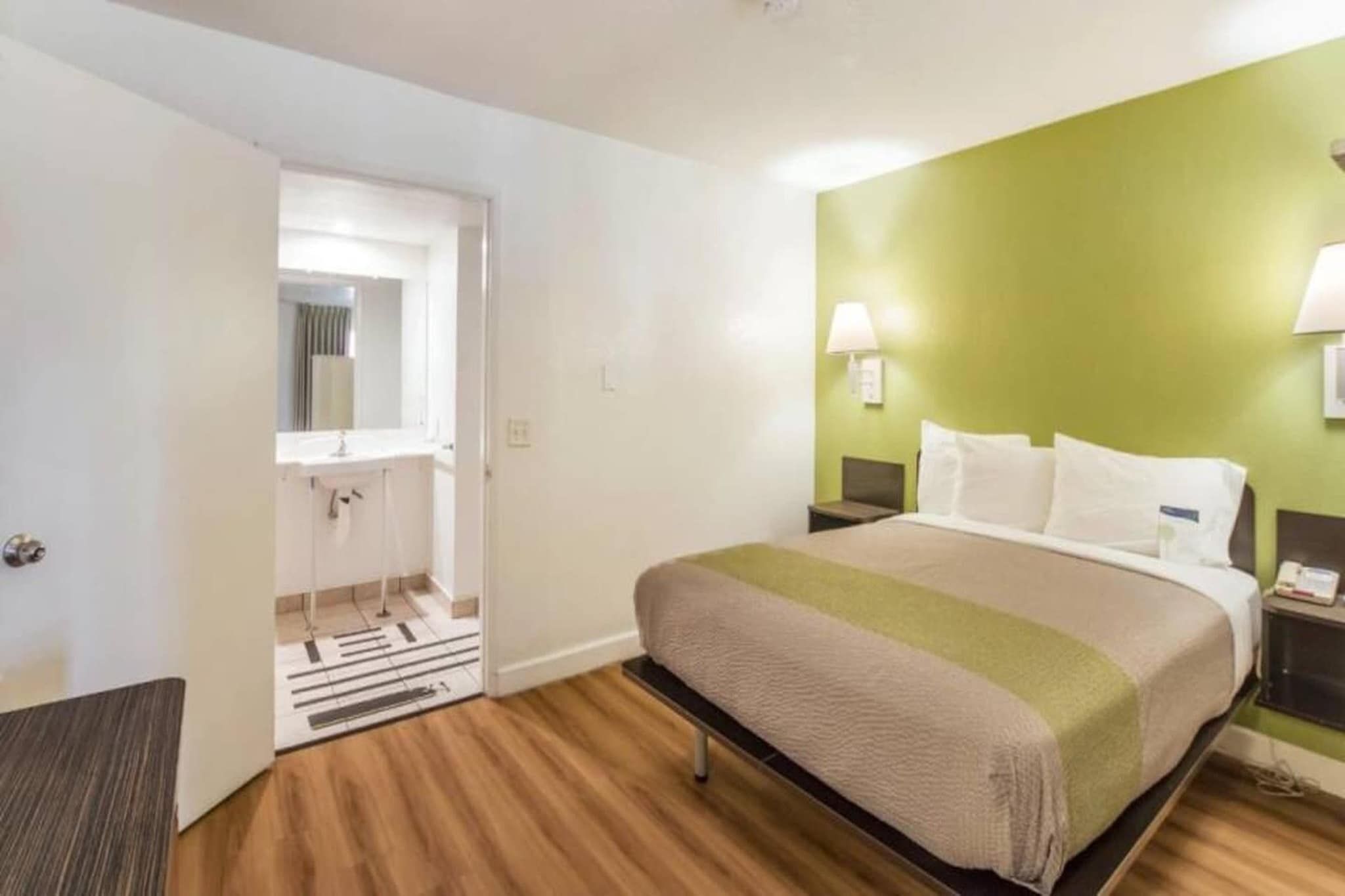 Hotellihuone, jossa on sänky, yöpöytä, lamppu, peilillä varustettu kylpyhuone ja ovi.