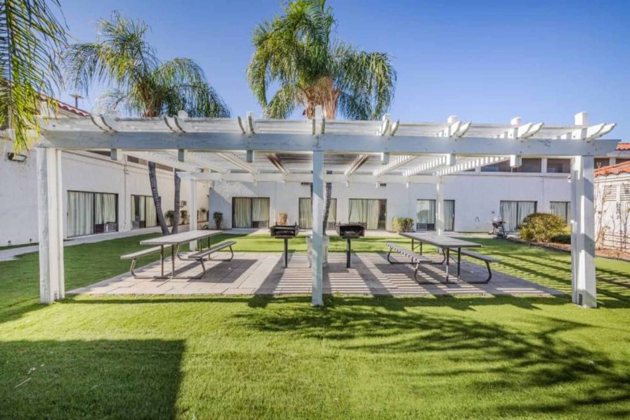 Un espacio al aire libre techado con mesas y sillas, rodeado por una pérgola blanca y palmeras al fondo.