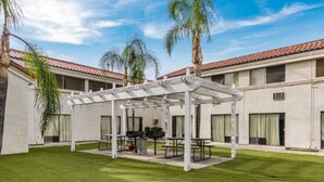 A courtyard with a covered seating area, palm trees, and a building with a tiled roof. - 1 Queen Bed | Non-Smoking (HEMET)