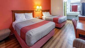 Quarto de hotel com duas camas, cabeceira de madeira, mesa de cabeceira com abajur, uma pequena mesa e uma cadeira.