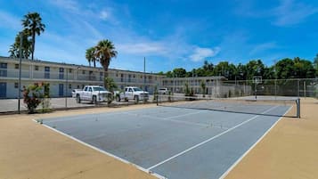 Una cancha de tenis al aire libre con red, rodeada por una cerca y contigua a un edificio de varios pisos con vehículos estacionados.